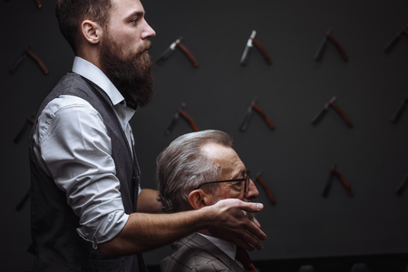 Bearded old businessman sitting at barber shop in chair choosing haircut designの写真素材