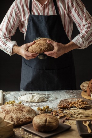male Baker in black apron holding freshly baked organic bread with a variety of bread, sour dough on foregroundの写真素材