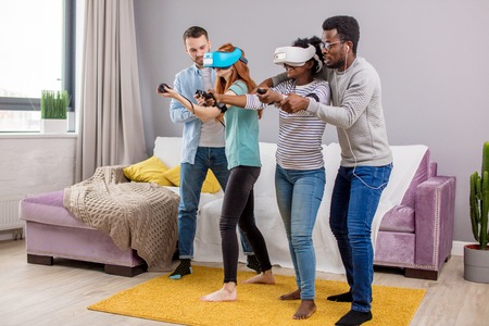 Multicultural group of friends playing video games wearing virtual reality glasses with controllers, women trying to play and boyfriends making hintsの写真素材