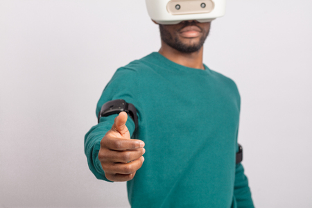 African guy in green poolover experiencing virtual reality using 3d headset for smart phone, stretching out hand with open palm as if inviting us to join himの写真素材
