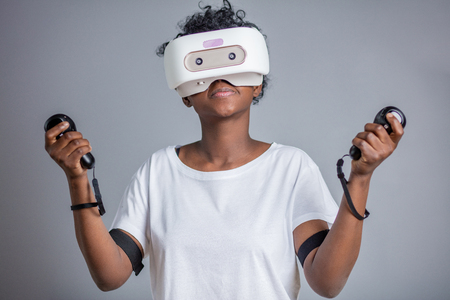 Beautiful young african woman in white t-shirt wearing white virtual reality headset and holds joysticks, posing ar camera over grey background.の写真素材
