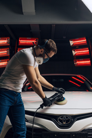 young man in mask using an auto tool after painting the car. close up side view photoの写真素材