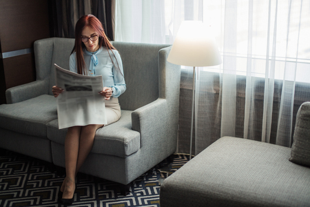Morning reading. Intelligent red-haired beautiful woman of european ethenity, dressed in romantic silk blouse relaxing in the armchair while reading newspapers in hotel room.の写真素材