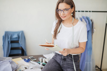 Young hard working seamstress with long blonde hair and spectacles sitting at her studio workplace...