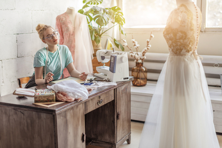Dreamy thoughtful seamstress with blonde hairmaking notes in sewing studio in soft pink and mint tones, making a work plan for the next weekの写真素材