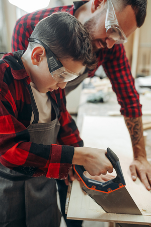 Caucasian young father in protective eyewear teaching his attentive pensive junior school son to saw wood in wooden workshopの写真素材