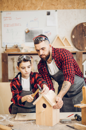 Young caucasian father and his pre-teen son working together in a wooden workshop, building a wooden bird house. Little boy dressed in apron and wearing protective glasses working with a hammerの写真素材