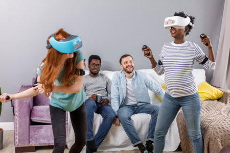 Group of young multicultural diverse friends using virtual reality headsets having fun at college campus. African girl and caucasian red-haired woman in VR glasses making virtual tour in shopping mallの写真素材