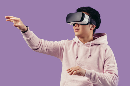Korean young man surprised by something in virtual reality, wearing virtual reality glasses showing gesture isolated on studio shot on violet wall backgroundの写真素材
