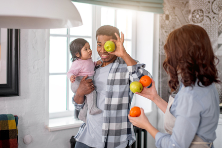 cheerful daddy having fun with fruit at home, happy guy with a kid in hand holding an apple in front of his eyes, happinessの写真素材
