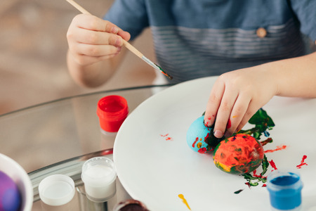 Home made Easter decorations and little helper. Selective focus of small hands of a boy covering boiled eggs with colourful paints on white plateの写真素材
