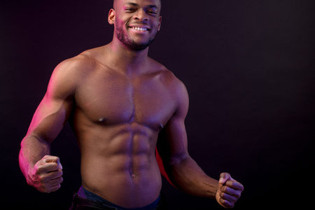 Attractive African male fighter or boxer posing shirtless, looking at camera, isolated over dark...