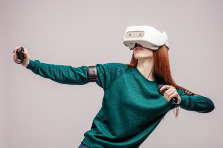 Red-haired caucasian young woman dressed in green casual sweatshirt, hanging head mounted display on head, aiming from bow with arrow, isolated over grey backgroundの写真素材