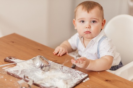Serious approach. Little helper with cute adorable eyes looking at camera while playing at kitchen. Cute little toddler boy crawling on kitchen table, covered with flour after making cakes.の写真素材