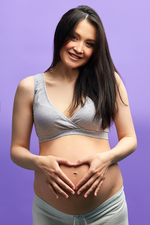 Pregnant woman in third trimester, dressed in comfotable home wear, posing against studio purple wall, looks happily at camera, being in anticipation of baby. Happy pregnancy and motherhood conceptの写真素材