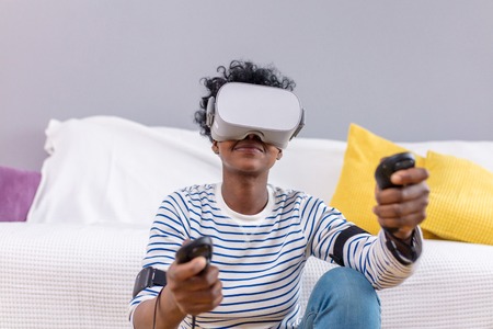 Indoor shot of pretty african female gamer using augmented reality headset with modern 3d mobile app.Young female experience virtual reality with smartphone and VR glasses on her head.の写真素材