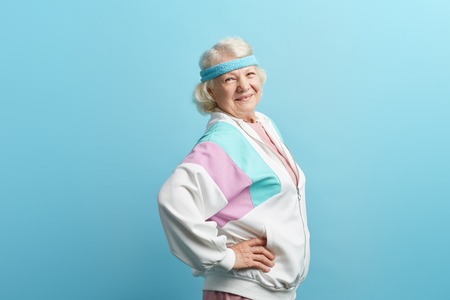 European retired woman in sportive clothings and headband, having beaming with joy face expression stands isolated in studio over blue background, copy spaceの写真素材