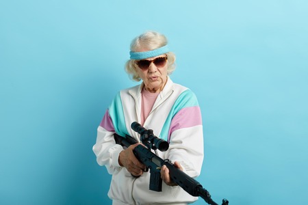 Experienced female agent working undercover of a grandmother-dandelion is pointing a rifle at the camera, standing isolated on blue background. Poster. Blue background.の写真素材