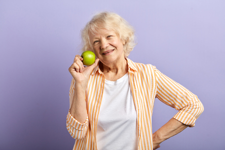Happy smiling mature woman in her sixties with trendy stripped shirt holding green apple and smiling at camera, isolated over purple backgroundの写真素材