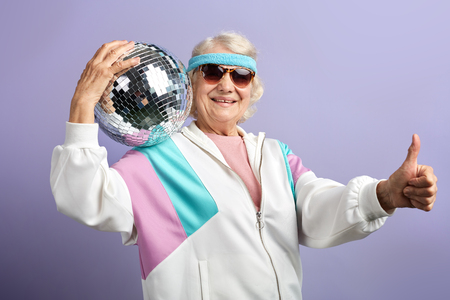 Joyful smiling aged woman portrait dressed in sportswear, holding ball, set in purple studio. Age of health, leisure, entertainment in retirementの写真素材