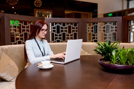 Attractive red-haired female blogger with blank badge has a video conference over the Internet  while sitting in restaurant and drinking coffeeの写真素材