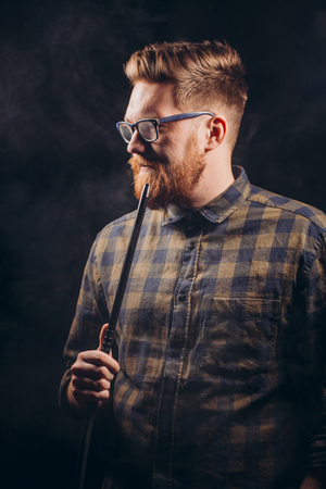 Stylish beard man in glasses and checked shirt smoking hookah at night bar.close eup shot. isolated black background. studio shot. free time. lifestyleの写真素材