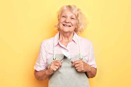 happy positive woman in grey apron with crossed spoon and fork looking at camera. isolated yellow...