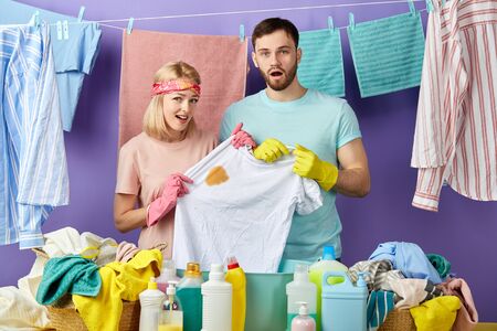 sad angry blonde woman in pink T-shirt and handsome man in blue T-shirt showing a spot on clothes to the camera. isolated blue background. washed clothes hanging on the clothesline. housework conceptの写真素材