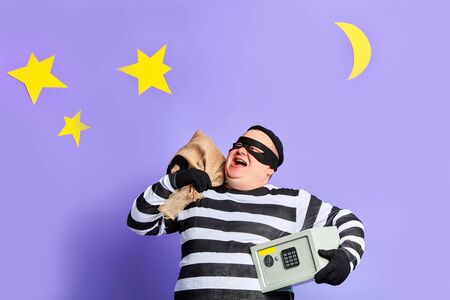 Happy cheerful plump robber carries sack and safe full of dollars . looking up, being happy to have a good criminal day isolated on blue.isolated blue backgroun with moon and starsの写真素材