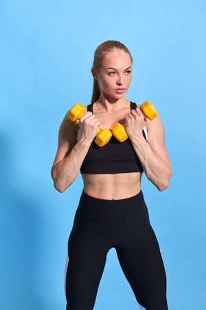 Beautiful muscular young woman in stylish sportswaer lifting dumbbells on blue background....