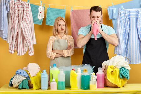 funny young man blowing to glove while blonde woman standing near him with crossed arms and looking at camera, yellow. studio shot. wet clothes hanging on clothesline in background of photo.の写真素材
