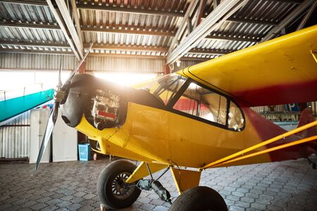 close up view of yellow single-engine propeller airplane standing in hangar building with opened motor cabinet, waiting for maintenance and service works.の写真素材