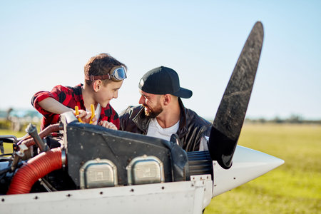 young cheerful man and smiling little kid doing routine maintenance of small propeller airplane,...