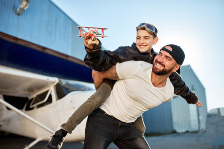 family portrait of son and dad playing together after the flight on their private small white airplane, outside aircraft hangar, boy holding toy plane, dressed in large pilot jacket.の写真素材