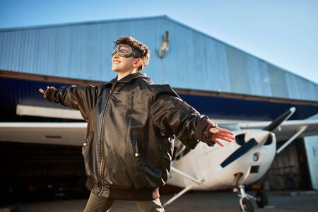 cute teenager boy holding hands apart like wings, standing outside hangar near single-engine air jet, waiting father to land at airfield, dressed in large fathers pilot jacket. Outdoor shot.の写真素材