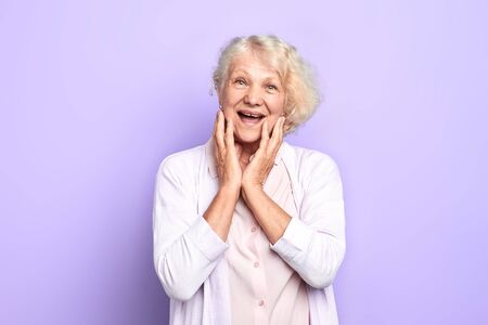 Old blonde girl touching face with palms, expresses happiness and joy. isolated blue background. studio shot woman has heard that she would get big pensionの写真素材