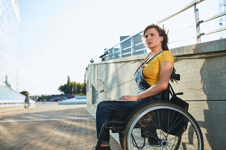 Disabled woman on wheelchair in T-shirt and overall using laptop outdoors.