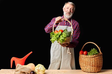 Friendly mature farmer shows results of hard work at his organic field and greenhouses, feels proud of rich havest, holds wicker basket with fresh raw vegetables. Studio shot, front view. Friendly mature farmer shows results of hard work at his organic field and greenhouses, feels proud of rich havest, holds wicker basket with fresh raw vegetables. Studio shot, front view.