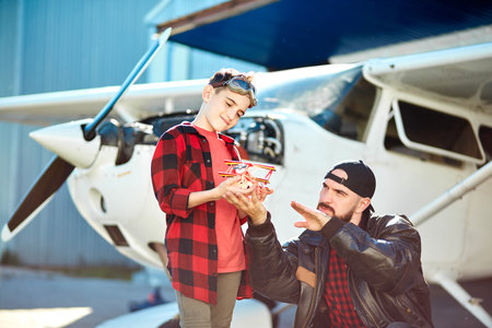 young handsome dad teaches and explains his kid aeronautical basics and principles, shows on toy plane flight routes, stand near light single-engine aircraft. Outdoor shot, aircraft hobby concept.の写真素材