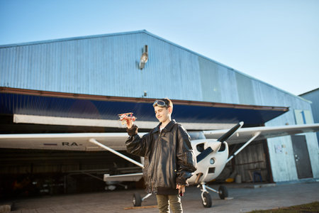 cute kid standing with smile, playing with small model airplane, wears aviator glasses and large...