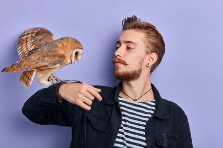 handsome young man is holding an owl in his arm, looking at it, teaching it to perform some actions. isolated blue background, job, profession, occupationの写真素材