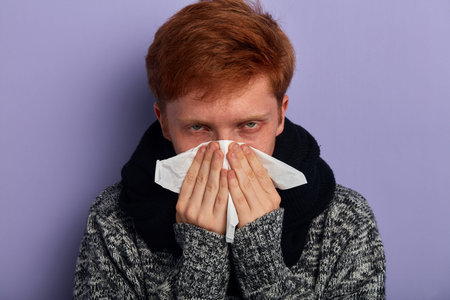 young unhappy serious man covers his nose part of the face by paper towel or tissue apparently...