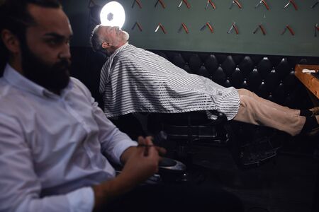 relaxed old man lying on the chair at barbershop, waiting for shaving, pensive serious killer in the foreground of the photo.の写真素材