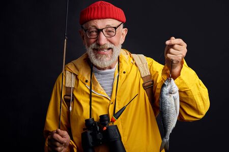 old experienced happy fisherman in glasses holding a fish and a fishing rod isolated on black background. close up photo. studio shot. hobby, interetsの写真素材