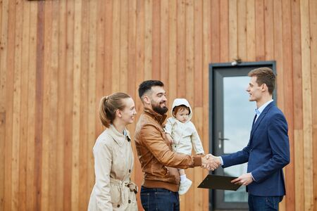 Caucasian happy family and realtor wear blue suit stand outdoor near new house just sold. Men shake hands.の写真素材
