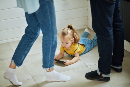 Caucasian child wear yellow T-shirt and denim overalls lies on the floor and plays on phone. Parents stand near in white and black socksの写真素材