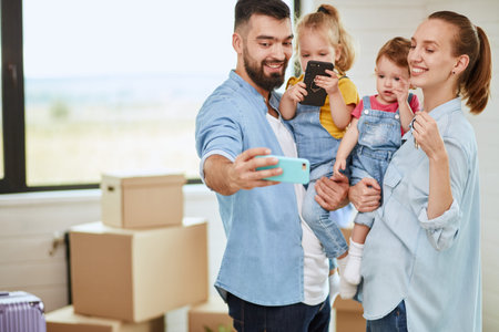 Caucasian family of four move in new house. Dad make photo. Background moving boxes, suitcase,...