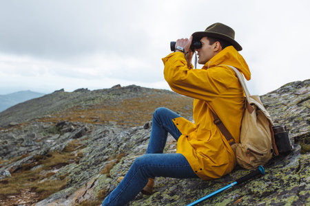 stylish hikers in raincoat, green hat with backpack holding binoculars sitting on top of the...