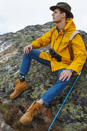 young hiker in stylish raincaot, green hat has climbed to the summit, sitting on it, close up side...