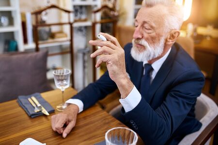 Elegant senior man in suite smoke heat-not-burn tobacco product technology sit in restaurant, holding cigarette in right handの写真素材